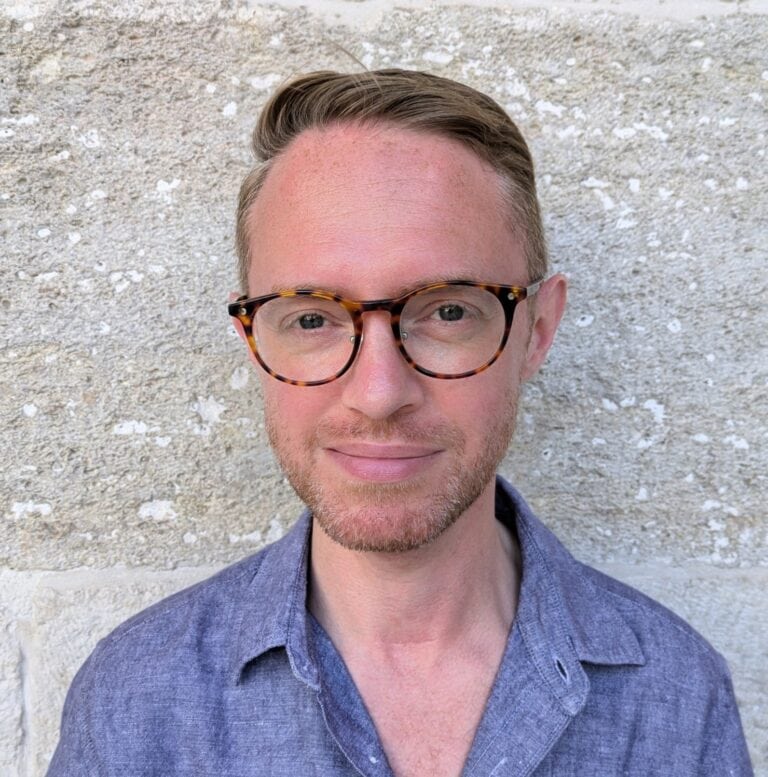 Portrait of a young man wearing tortoiseshell glasses and a blue shirt, standing outdoors against a textured stone wall, showcasing modern eyewear and fashionable casual style.